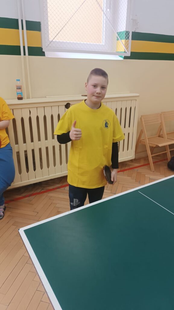 Mikołaj M. stoi uśmiechnięty na sali gimnastycznej w żółtej koszulce przed stołem do tenisa stołowego z kciukiem uniesionym do góry po wygranym meczu.