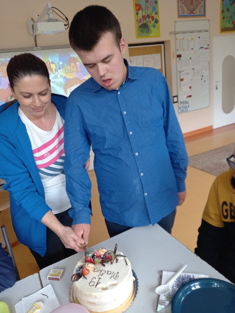 Chłopiec z pomocą nauczyciela kroi tort urodzinowy.