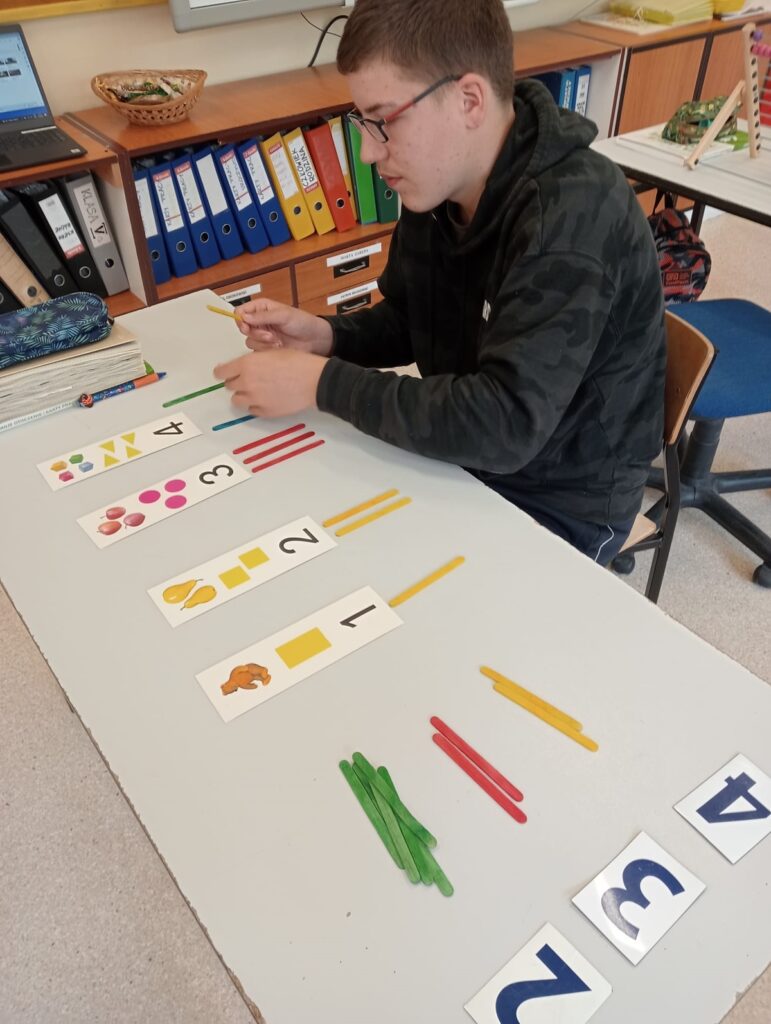 Uczeń układa kolorowe patyczki pod kartonikiem z cyfrą.